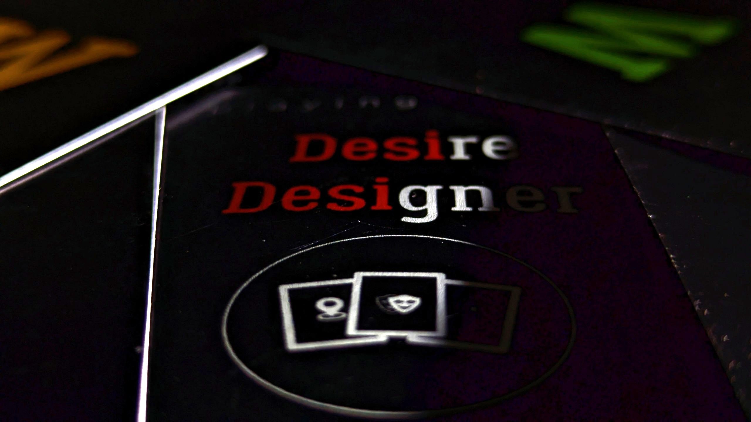 A close-up of a glossy black card deck box or packaging with the words 'Desire Designer TM' in red and white, plus a small icon of three cards inside a circular frame.